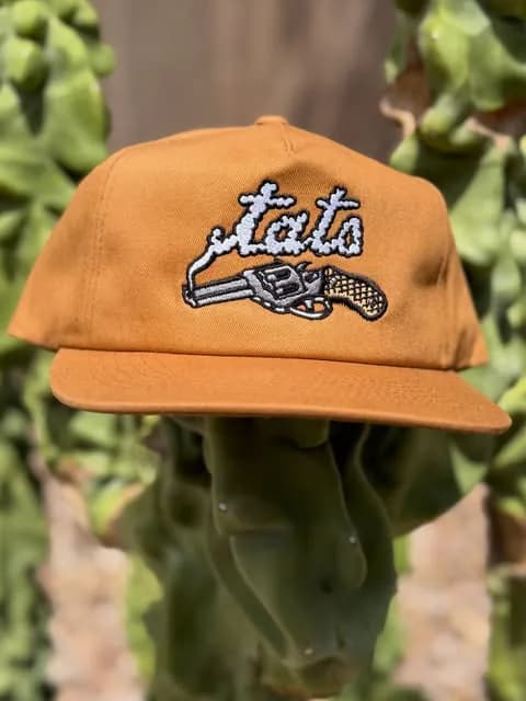 A rust-colored cap embroidered with a white revolver and Tats lettering