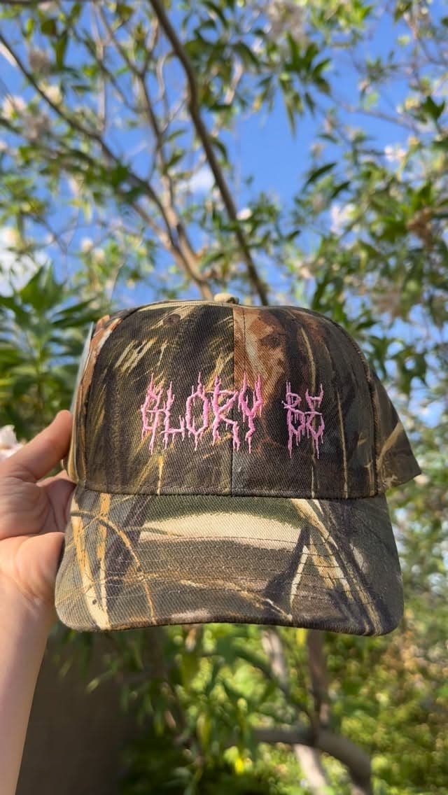 A camouflage cap embroidered with fine pink lettering held outdoors